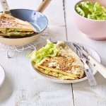 Een krokante tortilla ligt in een bordje klaar om aan te vallen. Hij is gevuld met een omelet, passendale kaas, gandaham en lente-ui. De tweede quesadilla ligt nog in de pan en staat ook op de tafel. 