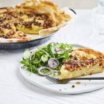 Er staat een quiche op tafel die aangesneden is. De quiche is gemaakt van schijfjes aardappel, ui, spek en rozemarijn. Wij serveren hem met een fris slaatje van kruiden op het bord, met een zoetzure dressing. 
