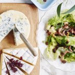 Een heerlijk salade met little gems, frisse appelstukjes en krokante spekblokjes staan op tafel in een glazen slakom. De slakom staat naast een kaasplank met een stuk blauwekaas en Belgische brie.