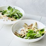 Salade met gegrilde asperges in 2 diepe borden. De groene salade bestaat uit veldsla en rucola met daarbovenop gegrilde witte asperges. Er is ook een yoghurtdressing met verse kruiden die bovenaan het bord staat in een glazen karafje. 