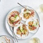 Sint-jakobsvruchten op een groot bord, opgediend in hun eigen Sint-jakobsschelp met bloemkoolmousse, witloof en spekjes. Glaasjes wijn en bestek ligt mee naast het bord, klaar om het feestmaal aan te vallen.