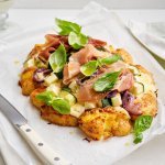 Een aardappelpizza ligt klaar om aan te snijden, en wat voor. Het is er eentje met een bodem van smashed potatoes, belegd met zure room, coutgette en rode ui. En afgewerkt met gedroogde ham en basilicumblaadjes. 