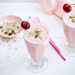 Twee rooskleurige smoothies op een witte houten tafel. De smoothie is afgewerkt met een kers en enkele pitjes en zaadjes. Een snel en makkelijk drankje met feestallures! 