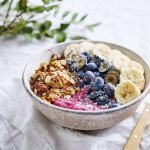 Een mooi gevulde ontbijtkom staat klaar. Onderaan zien we een dikke roze smoothie die als topping vers fruit kreeg, granola en kokosschilfers. Er ligt een lepel langs en servette om direct aan te vallen. 