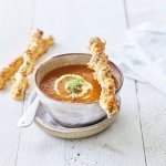 Een heerlijke kop soep staat te pronken op de eettafel. Het is een rood-oranje kleurige soep waarbij enkele bladerdeegstukjes liggen gevuld met kaas. De soep is afgewerkt met een lepeltje room en een peterselieblaadje. 