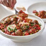 Op tafel staat een witte tajine heerlijk gevuld met gehaktballen in een rijke tomatensaus met eitjes. Er wordt net een portie uitgeschept. 