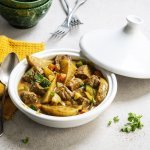 Op tafel staat een witte tajine met een gerechtje van rundvlees, courgette, selder, wortel en stukjes aardappel. Er ligt een okerkleurige servette bij en groene kommetjes om de tajine in op te scheppen. 
