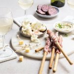 Een mooie marmere serveerplank in rond vorm staat op tafel. Hij is gevuld met allerlei tappas, zoals bloemkoolroosjes met cocktailsaus, wortel, kaasblokjes, salami met mosterd en homemade hummus.
