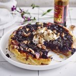 Een tarte tatin van rode kool met geitenkaas ligt op een groot wit bord. Er is al een stukje aangesneden en ze werd afgewerkt met enkele druppels balsamico en pecannoten. Achteraan zie je de fles balsamico nog op het aanrecht staan alsook een paars bloemetje. 