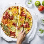 Een kleurrijke tomatentaart staat op tafel op een wit boterpapiertje. Ze wordt aangesneden door een onbekende vrouw met een koksmes. Rechtsboven liggen nog enkele tomaten om ze extra in de picture te zetten. 