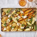 Ovenplaat met gegrilde groentjes komt net uit de oven en staat al op tafel. Hij heeft warme kleuren van de gouden aardappelen naar de oranje wortelen. Hij is afgewerkt met toefjes geitenkaas en verse chimichurri.