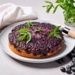 Op tafel staat een soort van tarte tatin, maar eentje met blauwe bessen en cakebeslag. Er ligt een mes langs om aan te snijden en enkele blaadjes verbena. 