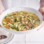 Een vegetarische minestrone met venkel, witte bonen, aardappel en verse kruiden geserveerd in een diep wit soepbord. Een vrouw met mooi rood gelakte nagels draagt het bord. 