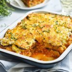 Er staat een grote schotel met vegetarische moussaka op tafel. Er is al een portie uitgeschept en op een bordje geserveerd. De moussaka is afgewerkt met verse tijmblaadjes. Op tafel staan ook nog 2 glazen en het plantje verse tijm.