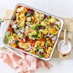 Op een witte tafel staat een houten snijplank met hierop een vierkante ovenschotel. Hij is gevuld met zoete aardappel, courgette, paprika en rode ui. Er is ook Berloumi gebruikt in dit gerechtje. 