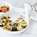 Vegetarische wok van aubergine en courgette staat klaar om aan te vallen in een witte wokpan. Er is een potje gevuld met tomatenblokjes, basilicum, azijn en olijfolie waarmee je je gerecht een extra frisse toets geeft.