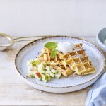 Wafeltjes van verloren brood liggen op een licht grijs bordje. De stukjes fruit van appel en peer liggen onder de wafeltjes. We gaven ook een toefje slagroom op de wafels voor extra smaak. Er ligt een blauwe servette en klein zeefje met bloemsuiker langs het bord.