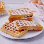 Een stappeltje klaargemaakte wafels ligt op een groot wit bord. Bloemsuiker is rijkelijk op de wafels gestrooid. In de achtergrond zie je nog enkele eitjes liggen en een roze linnen servette.