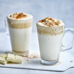 Twee hoge glazen tassen staan op het aanrecht klaar om te serveren. Ze zijn gevuld met witte chocolademelk, een goeie toef speculaasroom er bovenop en wat speculaaskruim. Er ligt een stukje witte chocolade naast. 