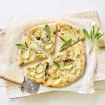 Een pizza belegt met mozzarella, plakjes aardappel en salie ligt op een wit boterpapiertje. Er ligt een pizzasnijder langs de pizza en nog enkele verse blaadjes salie. 