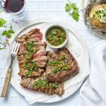 Het heerlijk stukje vlees ligt op een groot wit rond bord. Het al voor de helf in reepjes gesneden en bedruppeld met de Aziatische chimichurri. Er ligt een gepofte aardappel langs en twee glaasjes rode wijn. 