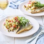 Op tafel staan twee borden met het gerechtje op. Het is een aardappelpuree met stukjes paprika, kappertjes en rucolasla. En een krokant gebakken zeebaarsfilet. Er staat ook een glaasje witte wijn langs. 