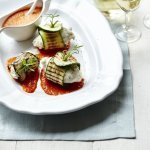 Op een grote witte schaal liggen meerdere rolletjes gemaakt van een stukje vis met gegrilde courgette of aubergine. Dit alles is afgewerkt met een coulis van paprika.