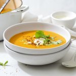 De soepkom staat op tafel en is gevuld met een prachtige warme oranje soep, nl zoete-aardappelsoep. De soep is geserveerd in mooie witte soepkommetjes en is afgewerkt met een lepeltje zure room, chilivlokken, lente-uitjes en koriander. 