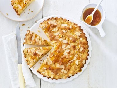 Goudgele appelcake staat op een houten witte tafel. De ronde cake is al voor een kwart aangesneden. Er is al een stukje uitgenomen en er staat een potje met abrikozengelei bij. 