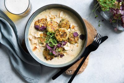 Balletjes van champignons en bonen met mosterdsaus Balletjes van champignons en bonen met mosterdsaus in een diep grijs bordje op een ruwe houten plankje. Linksboven staat een versgetapt bier en rechts nog een portie extra groenten. Onderaan ligt nog een grijze linnen servette en zwart bestek langs het bord.