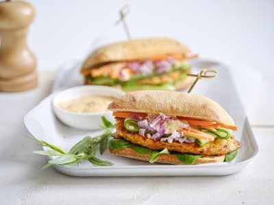 Een belegd broodje ligt op een bordje. Hij is belegd met een stukje gebakken kipfilet, verse wortel, komkommer en een sausje. De resterende saus werd in een klein potje erbij geserveerd.