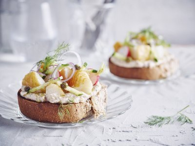Bruschetta met bonenpuree, gerookte heilbot, radijzen en komkommer Een goedgevulde bruschetta ligt op een klein glazen bordje en staat op tafel. De bruschetta werd afgewerkt met een takje dille. Achteraan de tafel staat een kan met water en bestek om deze toast te verorberen.