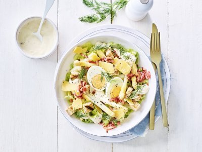 Caesar salade met asperges Een rijkgevulde salade staat klaar op tafel. Het witte bord is gevuld met sla, gekookte eitjes, stukjes asperges en kaasschilfers. Dat is de lente proeven in je bord, als je het mij vraagt!