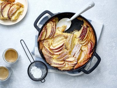 Clafoutis van appel Een clafoutis van appelstukkjes staat op het aanrecht in een zwarte fonte schaal. Er is al een portie uitgeschept en staat op een bordje naar de pot. Er is ook bloemsuiker over het dessert gestrooid en de koffie is al uitgeschonken.