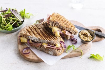 De croque ligt op een houten plankje aangesneden. De kaas en rode ui zien er heerlijk uit. In de achtergrond zie je ook nog wat gemengde sla en een potje graantjesmosterd. 