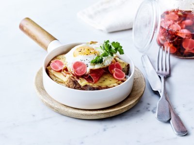 Croute au fromage met Averbode kaas in een witte pan, spiegeleitje er bovenop gepresenteerd met een takje verse peterselie. Op de achtergrond staan gepekelde radijzen in glazen pot.