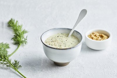Dillesaus met mosterd De dillesaus is geserveerd in klein porselein potje, er zit een witte lepel in en een takje dille langs.