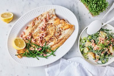 Gebakken schartong met zilte aardappelsalade Een heerlijk krokant visje afgewerkt met zeekraal ligt op een groot bord. Er zijn enkele garnaaltjes aan toegevoegd die mooi roze blinken. De aardappelsalade staat er langs in een witte kom met witte lepel. Om af te werken ligt er 1 citroen doormidden gesneden.