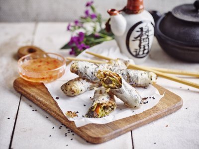Gebakken springrolls met buikspek uit de airfryer Heerlijke krokante springrolls liggen op een houten snijplank. Ze zijn vergezeld met een zoetpikante dipsaus. In de achtergrond staat nog een theepot en paarse bloemetjes.