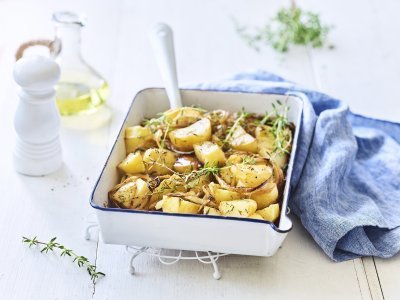 Een witte ovenschaal met donkerblauw randje staat op een keukenonderlegger op een witte houten tafel. Er staat een witte pepermolen en een glazen flesje met olijfolie langs. De verse tijm zorgt voor extra kleur bij de geroosterde aardappelen, er ligt ook nog een blauwe linnen servette langs de ovenschotel.