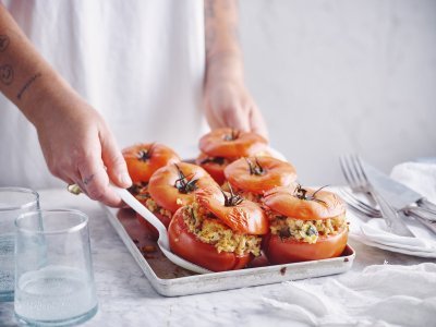 Zes gevulde rode tomaten in een ovenschaal zijn aan tafel gebracht door een vrouw in witte t-shirt. Ze schept alvast eentje op voor zichzelf.