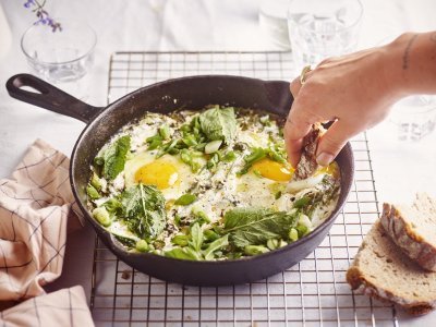 Groene shakshuka met spinazie, lente-uitjes, munt en yoghurt Op een rooster staat een zwarte gietijzeren pan met grieks getint gerechtje. Groene shakshuka met gebakken yoghurt. Er is een hand met een stukje brood in beeld dat er een dipje van zal nemen.