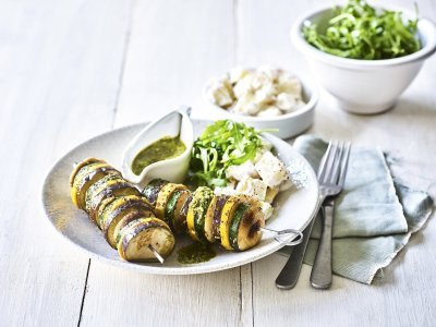 Op een bleke houten tafel staat een diep bord met hierin een schep aardappelsalade in, twee groentespiesjes met schijfjes van gegrilde aubergine, courgette, champignons en een kannetje met rucolapesto. Verder staat er ook nog een kom met verse groene salade, bestek en een muntkleurige servette.
