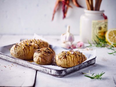 Klaargemaakte hasselback potatoes met pittige kruiden. Ze liggen op een grijze ovenschaal op de keukentafel. Achteraan het beeld zie je hete pepers, look, rozemarijn en een halve citroen liggen.