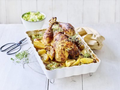 Hoevekip uit de oven met appeltjes en krieltjes De gebraden kip ligt te blinken in een ovenschaal met krieltjes en appeltjes naast. Het gerechtje is afgewerkt met groene verse tijmtakjes, het zwarte schaartje waarmee de kruiden werden afgeknipt ligt er langs.