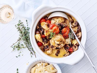 Stoofpotje van kip met tomaten en champignons. Op een wit tafelkleed staat de stoofpot geplaatst, hierin zit een grote lepel om de stoofpot uit te scheppen aan de gasten. Er staat linksboven een glaasje rosé in beeld en verse taktjes tijm.