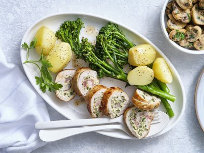 Kipfilet gevuld met een mengsel van roomkaas, mosterd, kruiden en noten. Om af te sluiten voegde we ook nog enkele lapjes spek toe en rolde we hem dicht. Hij ligt aangesneden op een ovaal bord vergezeld van gekookte aardappelen en gekookte broccolini. De gebakken champignons staan in een apart kommetje met een takje peterselie. 