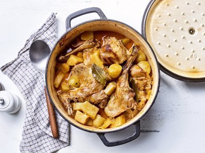 Konijnenragout met knolselder, pastinaak en aardappel Op tafel staat een gietijzeren pot gevuld met de ragout van konijn. Hij heeft een gouden kleur door de pastinaak, wortel en krieltjes.