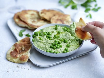 Krokant platbrood met erwtendip Krokant gebakken platbrood ligt op een serveerschaaltje waarbij een dip van erwten en roomkaas in geserveerd. De dip is gemixt en heeft een mooie groene kleur, om af te werken zijn er nog enkele verse erwtjes bovenop gestrooid. Een hand neem alvast een stukje platbrood om te dippen in gezonde groentedip.