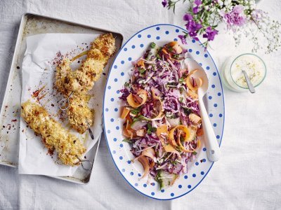 Krokante kipbrochette met een slaatje van kool, komkommer en wortel Krokante kipbrochette ligt nog de sidderen op een ovenplaatje. Daarnaast staat een kleurrijk groot bord met een salade op basis van rode kool, wortel en komkommer. De dressing op basis van yoghurt staat langs de salade.